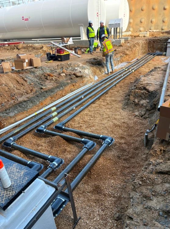 Construction workers in safety gear inspecting underground piping at an industrial site with large tanks nearby.