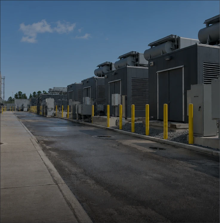 Row of industrial buildings with yellow safety bollards along a paved road under a blue sky.