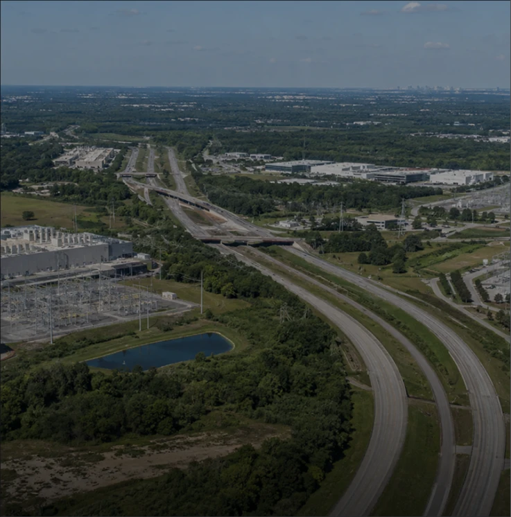 Aerial view of a curving empty highway surrounded by trees, industrial buildings, and a pond.