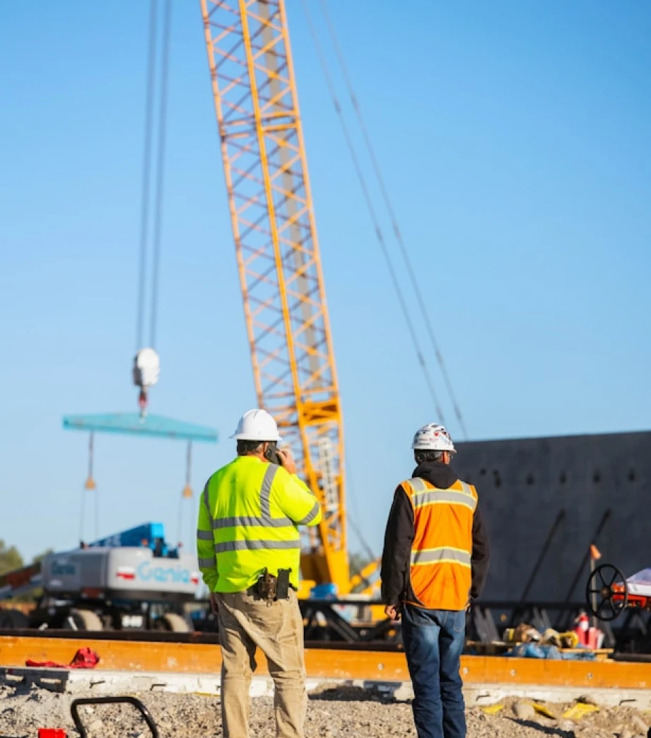 Two construction workers wearing safety gear observing work at an active construction site with a crane.