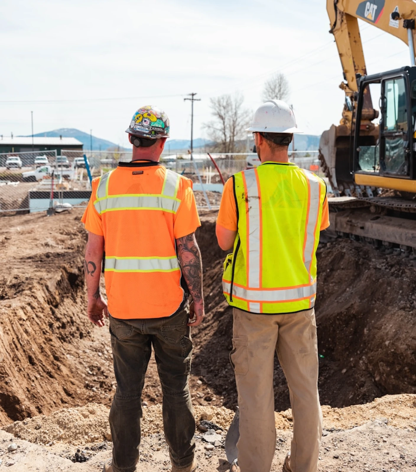 Two construction workers in safety vests and helmets standing near an excavated site with machinery.