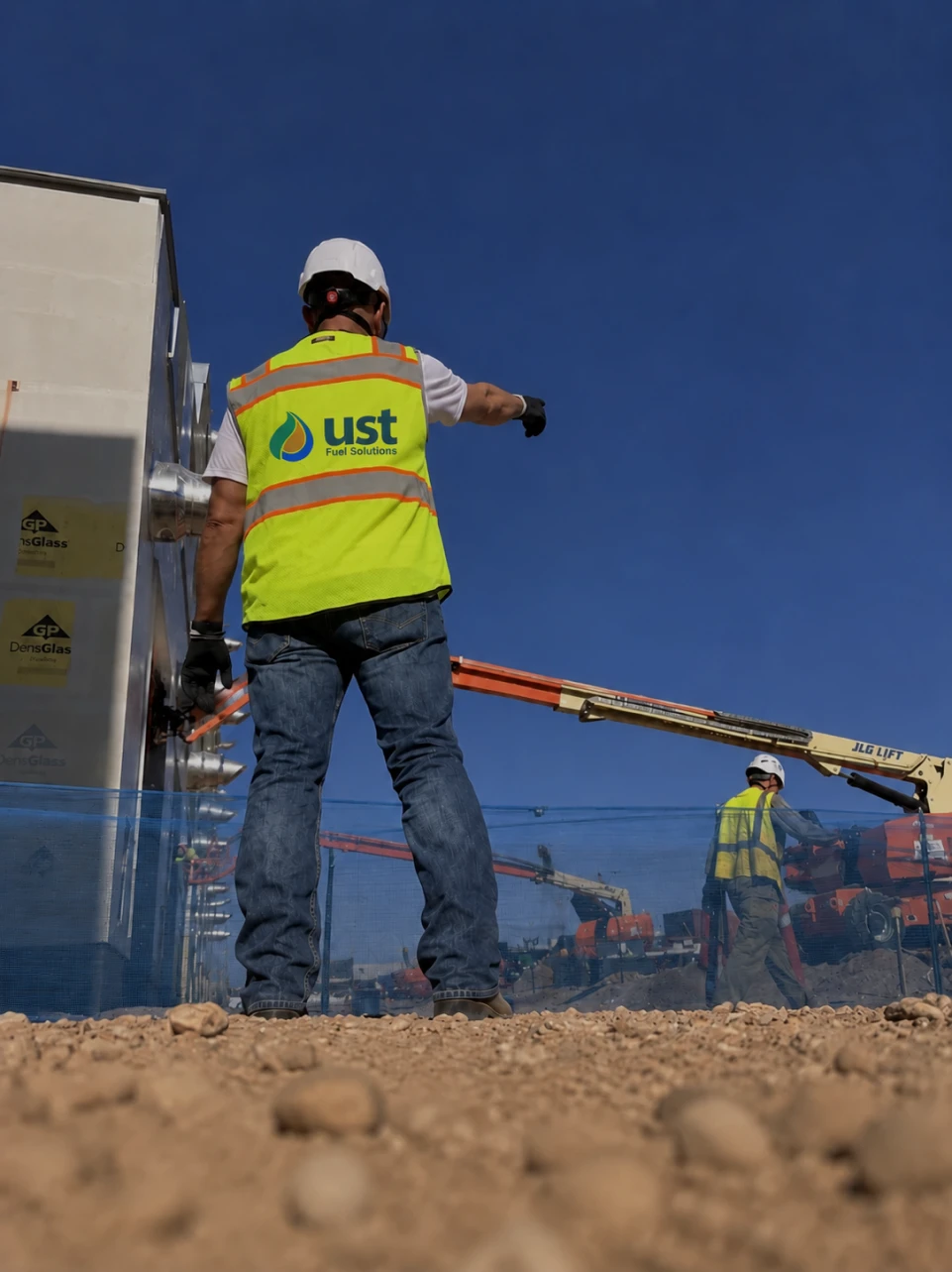 Construction worker in a hard hat and yellow vest pointing on a site with machinery under a blue sky.