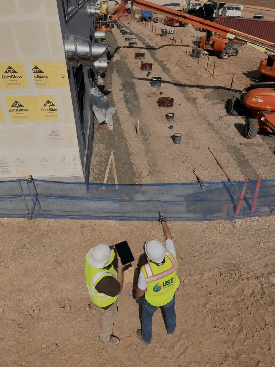 Two construction workers in safety vests and helmets reviewing plans at a construction site with equipment.