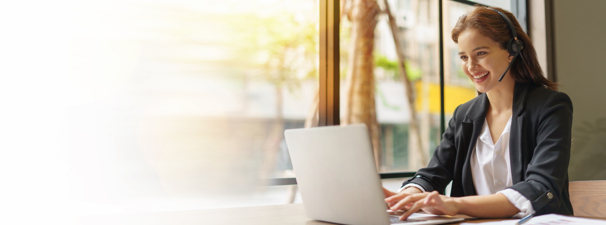 Smiling woman wearing a headset and black blazer, working on a laptop at a desk near a large window with sunlight and blurred trees outside.