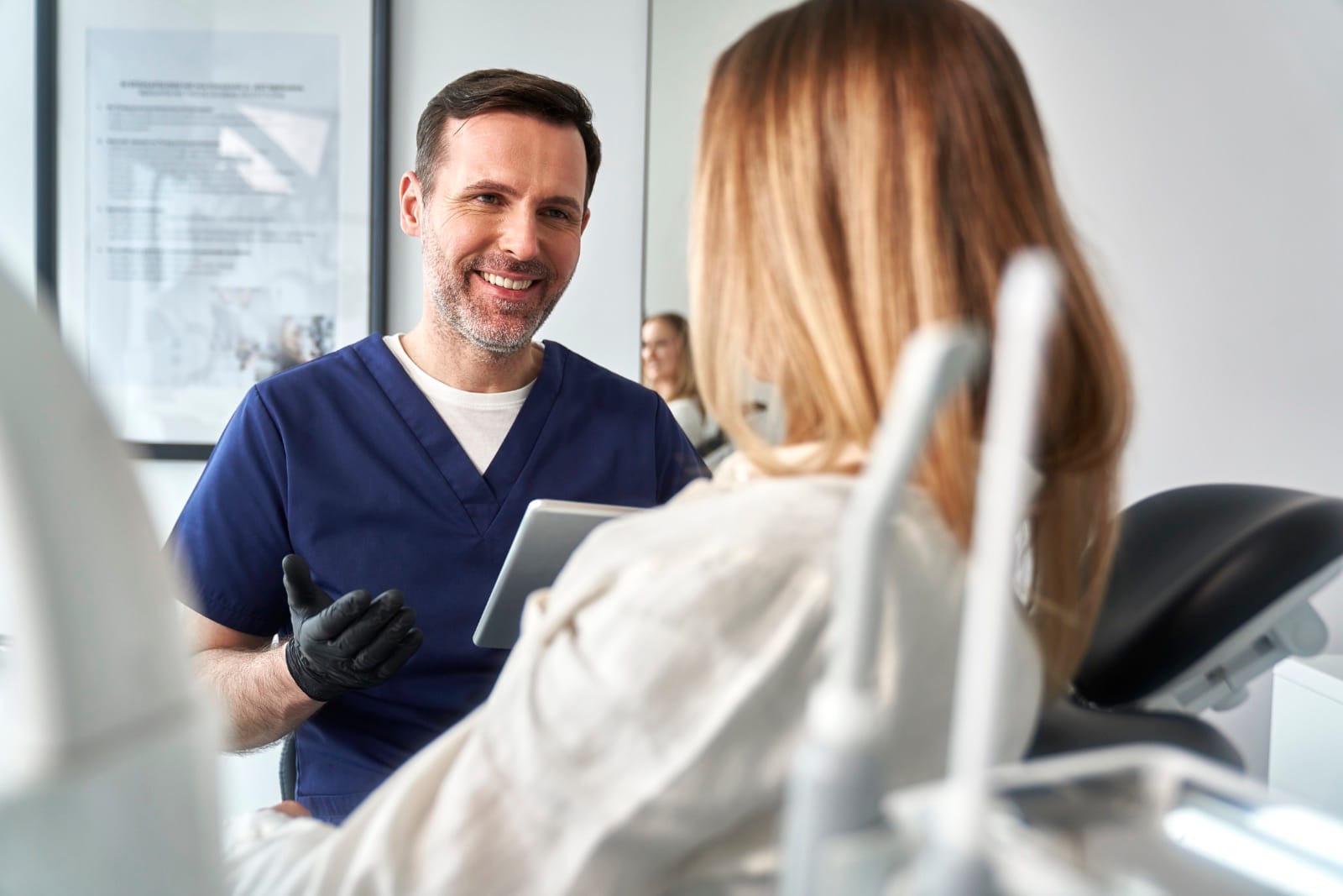 Male dentist in navy scrubs wearing black gloves smiling and talking to a female patient seated in a dental chair, holding a tablet in a bright dental office.