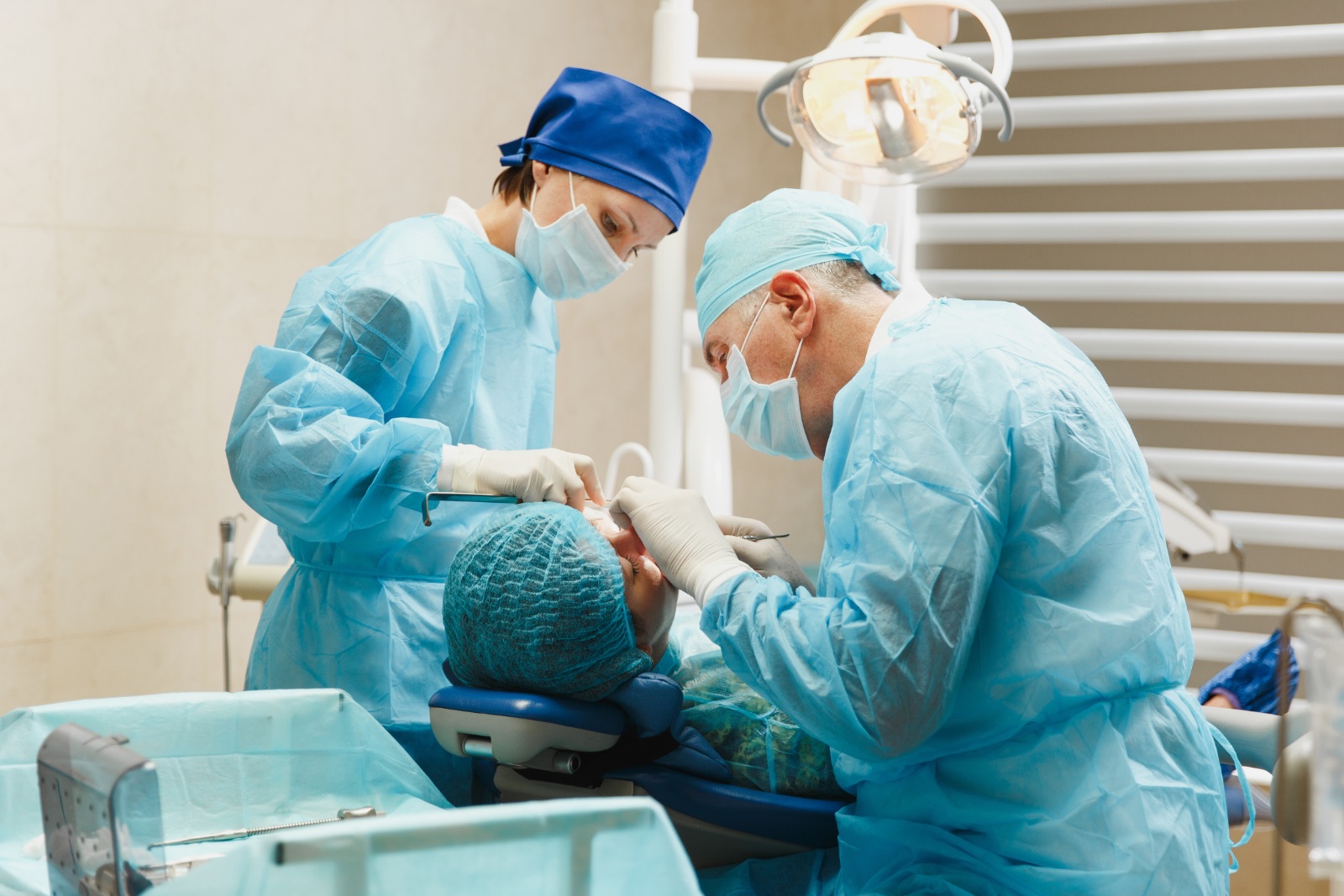 Two dental professionals in blue scrubs, masks, and hair covers perform oral surgery on a patient lying in a dental chair under an overhead light in a clinic.