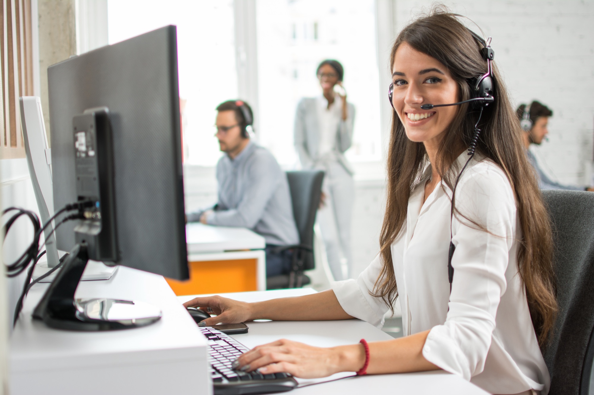 Smiling woman wearing a headset sitting at a desk in a call center or office environment with colleagues in the background also wearing headsets and working at computers.