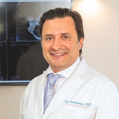 Smiling man in a white dental coat with 'Ilya Benjamin, DMD Sedation Dentistry' embroidered on it, standing indoors in front of an X-ray image.