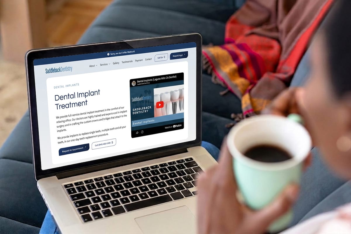 Person holding a cup of coffee while viewing a dental implant treatment webpage on a laptop, which includes text about implant services and a video about dental implants.