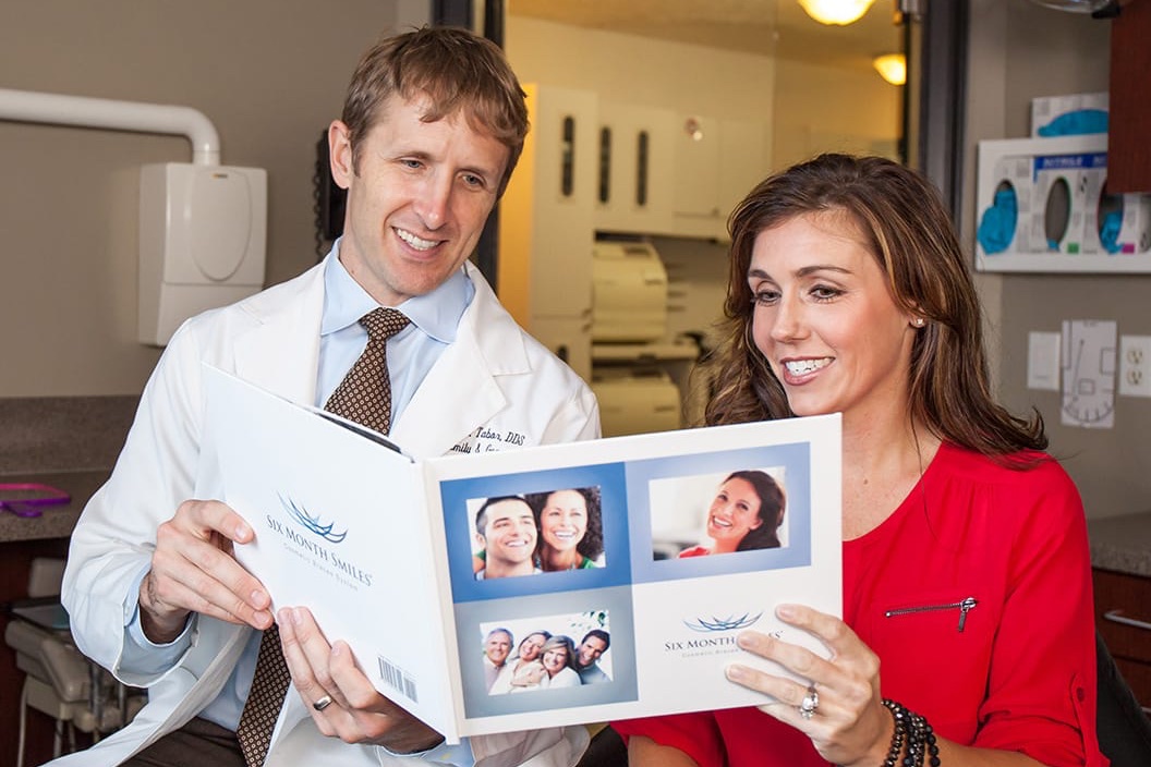 A male dentist in a white coat and a female patient in a red blouse are sitting together in a dental office, looking at and smiling over a brochure titled 'Six Month Smiles' featuring photos of smiling people.