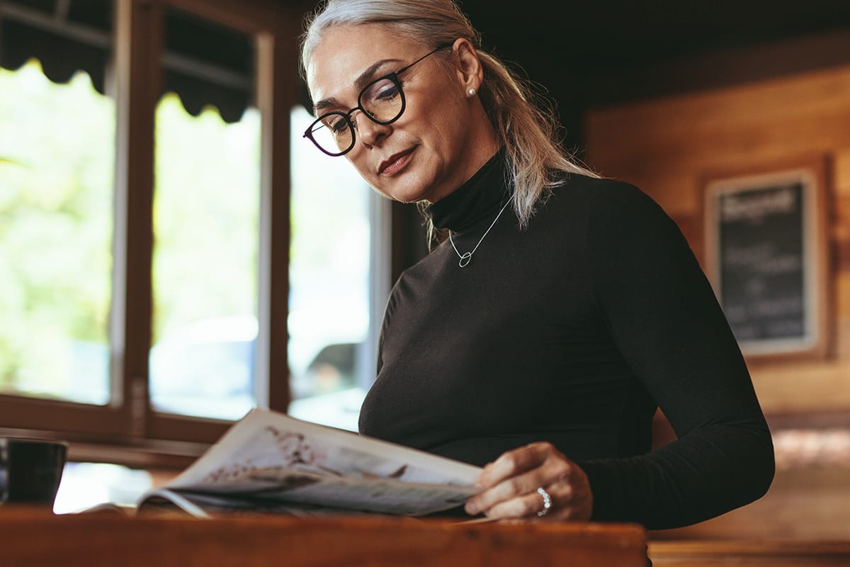 A middle-aged woman with gray hair and glasses reads a magazine while sitting at a wooden table in a cozy room with large windows and warm lighting.