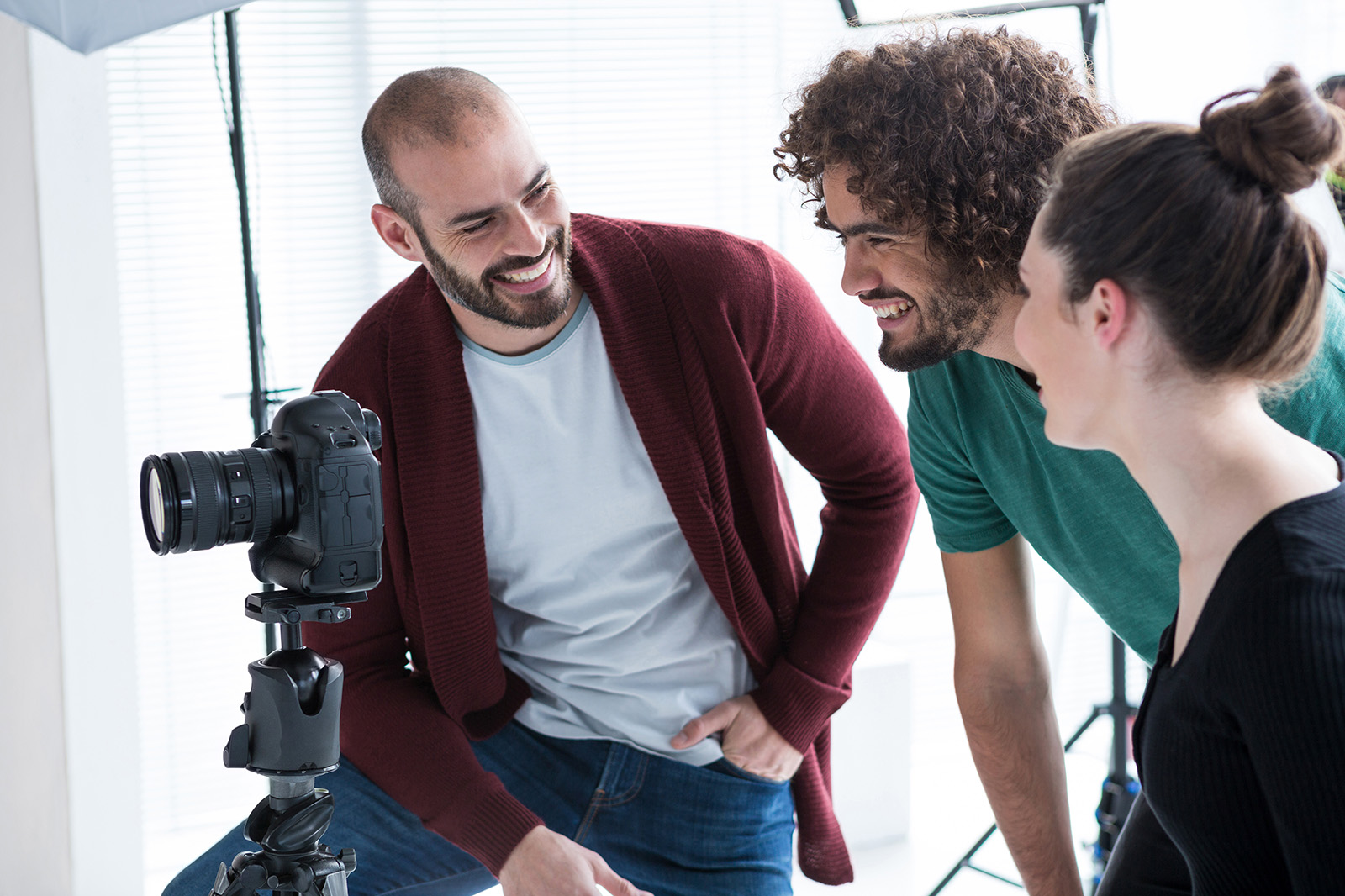 Three people smiling and looking at a camera on a tripod in a bright studio with photography equipment.