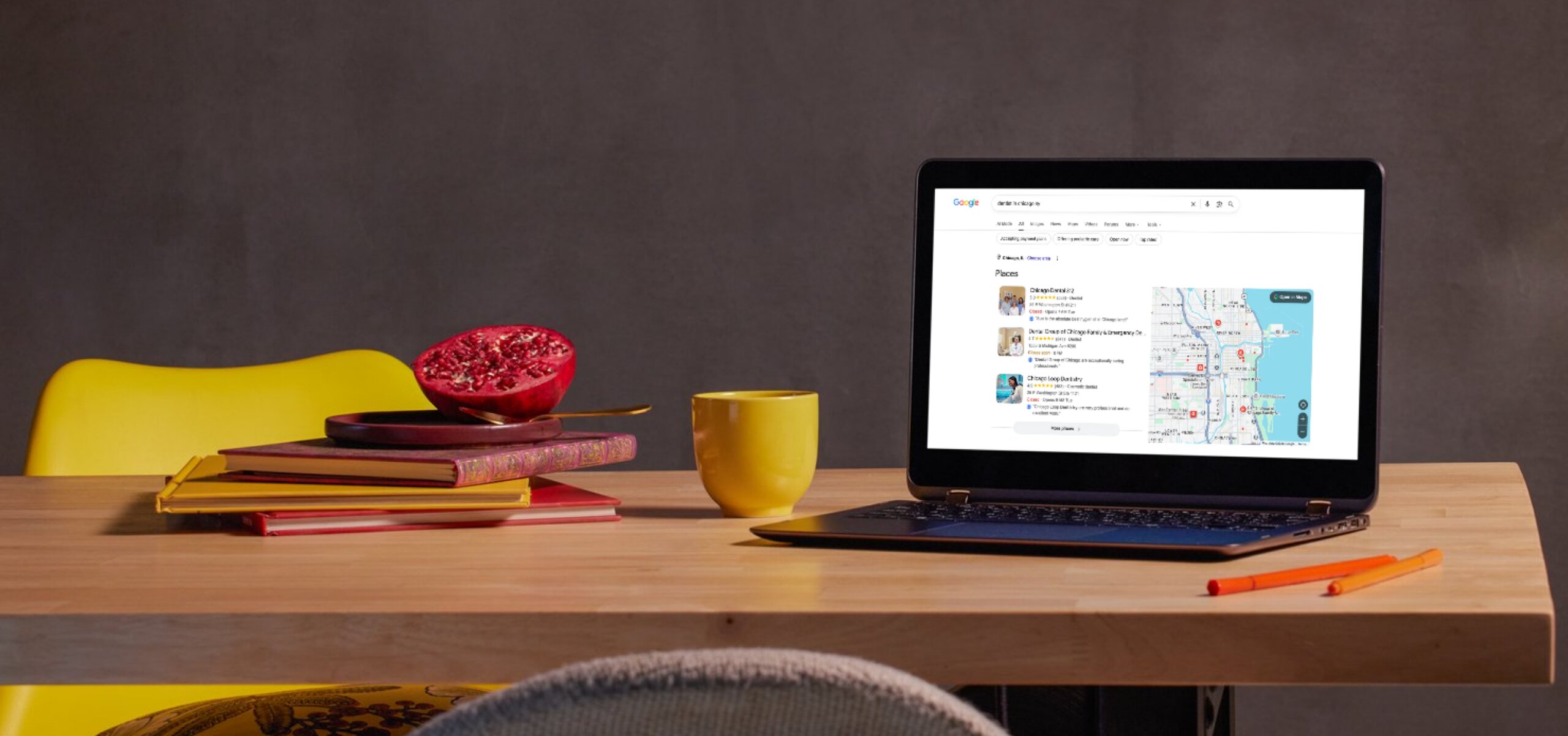 Wooden table with a laptop displaying a Google search for 'dentist in chicago il' showing local dentist listings and a map, a yellow mug, two orange pens, stacked books, a plate, and a halved pomegranate with seeds visible, with a yellow chair behind the table.