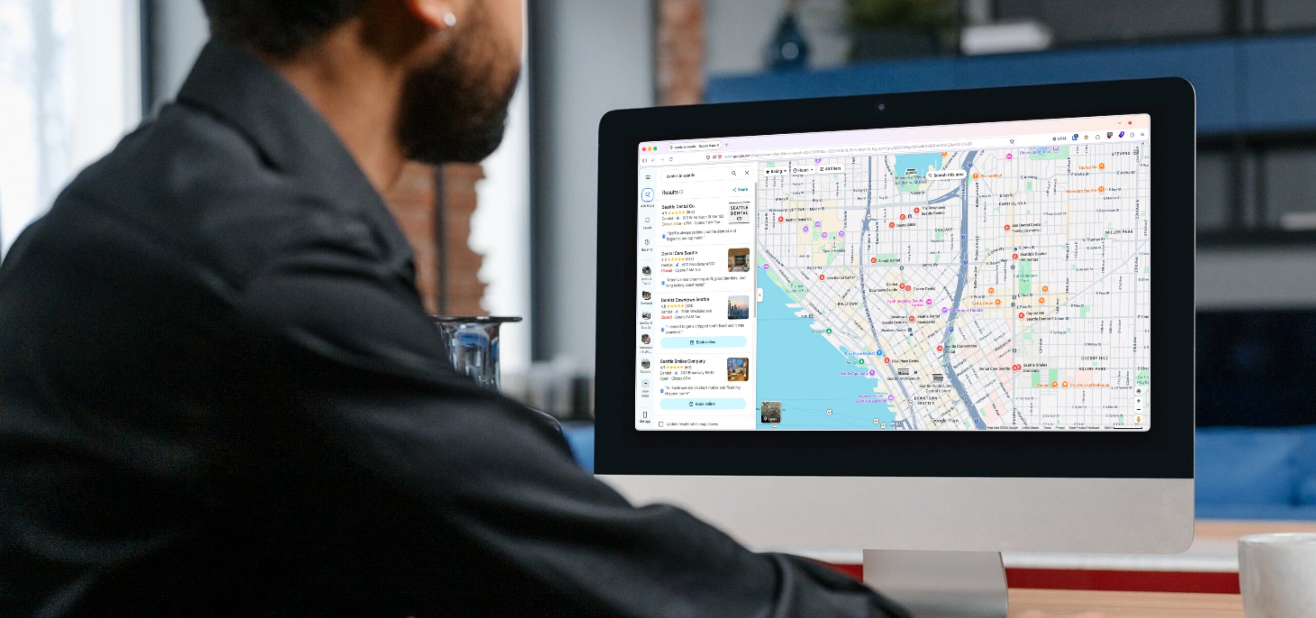 Man in a black shirt looking at an iMac screen displaying a map with location pins and business listings on the left side.