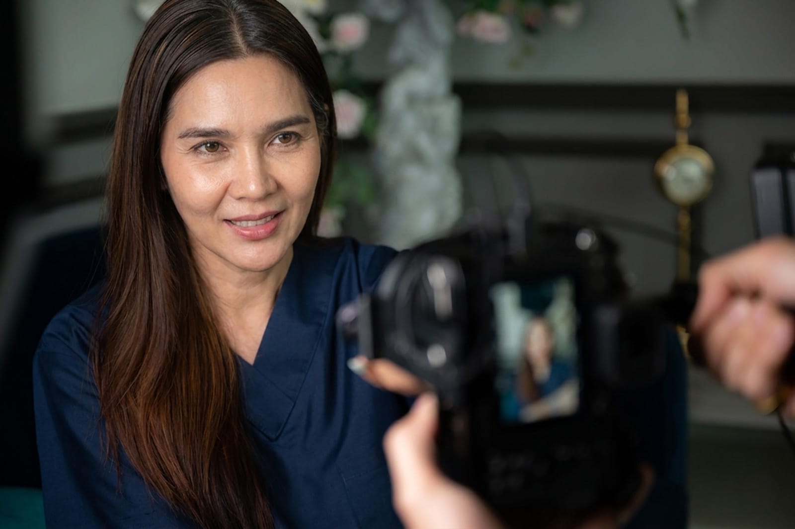 A woman with long dark hair wearing a dark blue top smiling while being recorded or photographed by a camera held by another person in a blurred background setting.