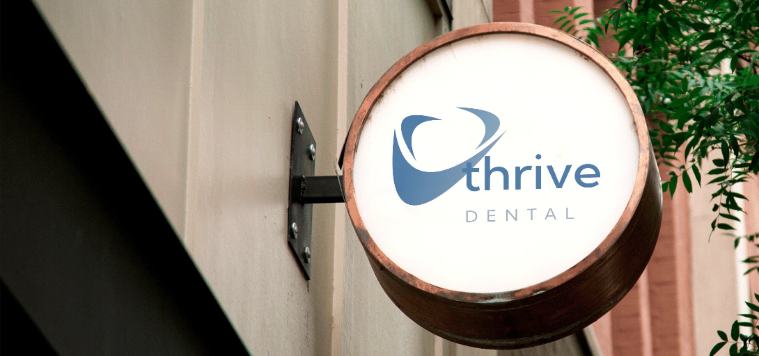 Round sign mounted on a wall with blue logo and text that reads Thrive Dental, with greenery in the background.