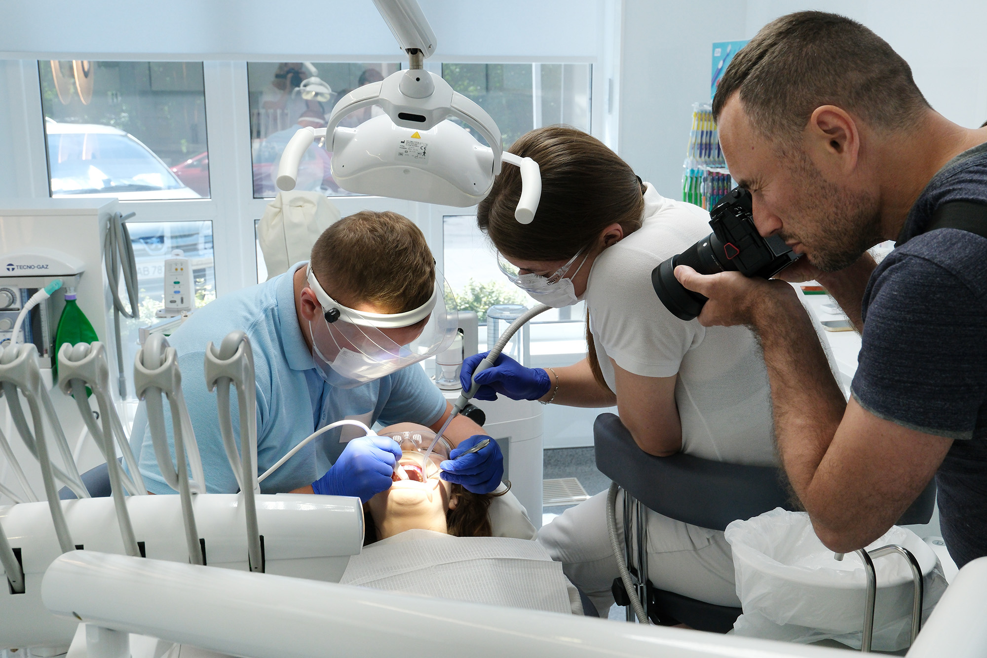 Dentist in blue scrubs and face shield treating a patient's teeth with assistance from a woman in white scrubs holding a suction tool, while a man with a camera photographs the procedure inside a bright dental clinic.