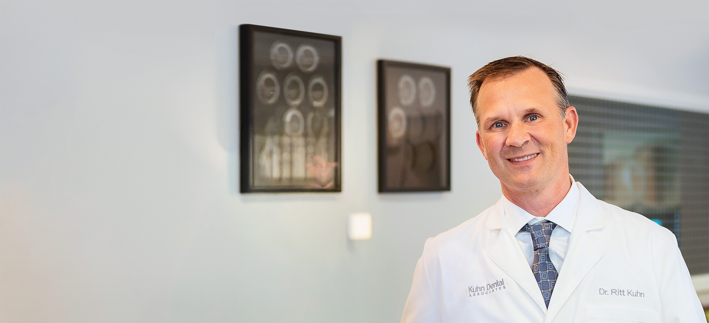 Smiling male dentist in a white coat with a patterned tie standing in a dental office with dental X-rays on the wall behind him.