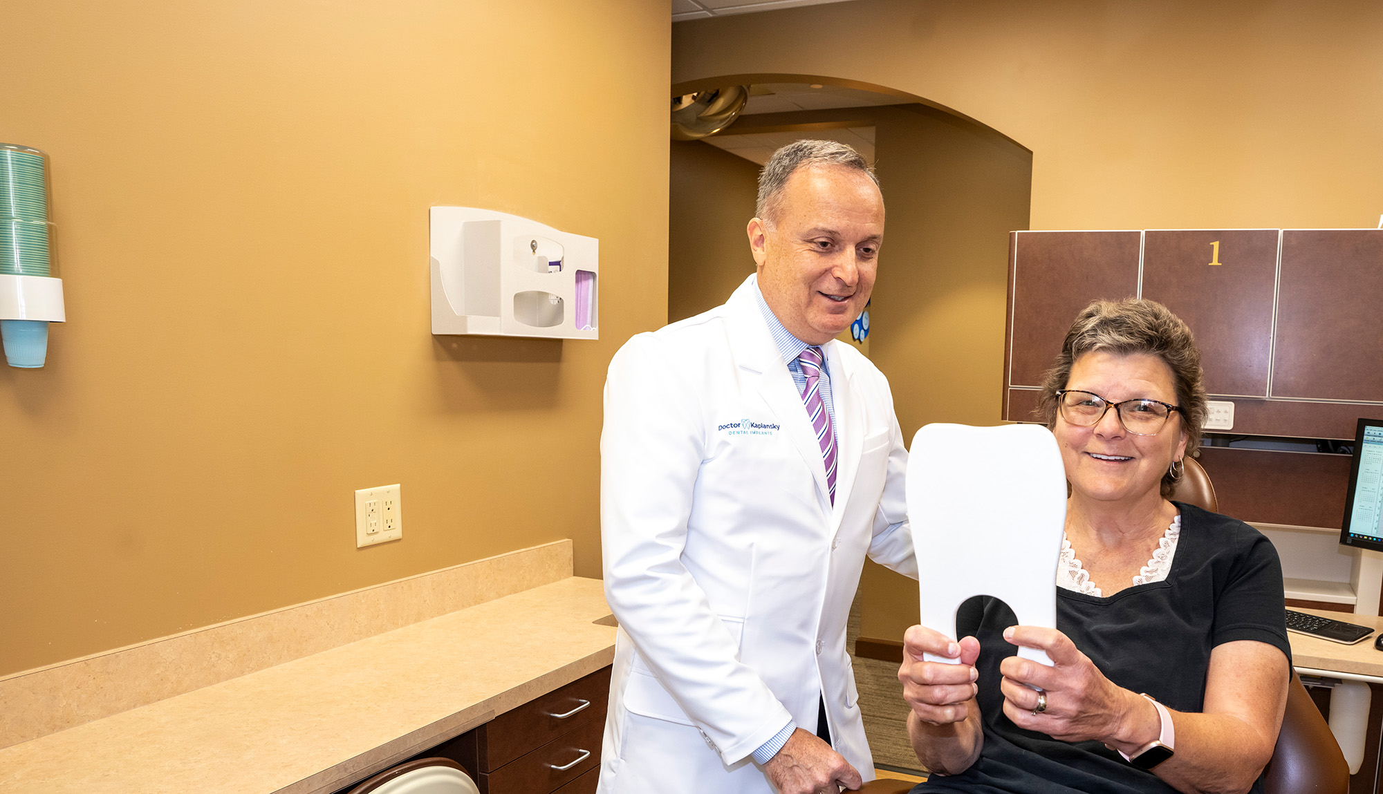 A dentist in a white coat standing next to a smiling woman seated in a dental chair holding a mirror in a dental office.