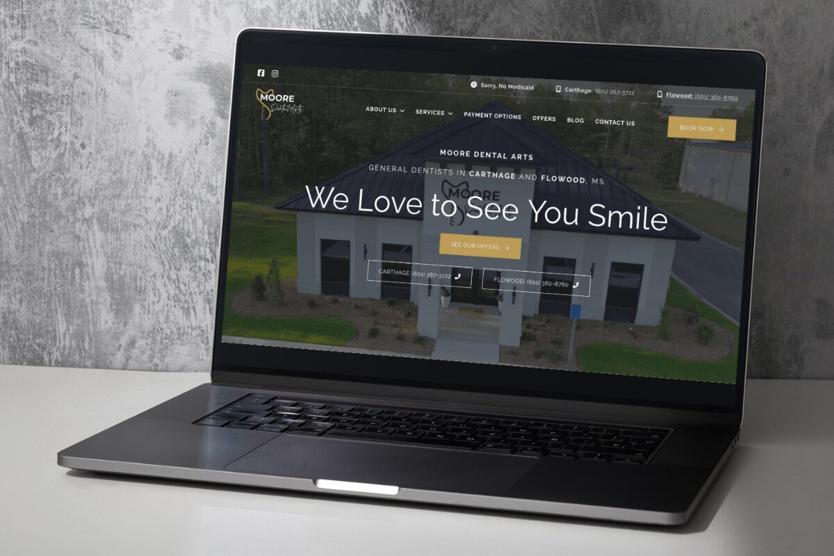 Laptop on a white surface displaying the Moore Dental Arts website homepage featuring their slogan 'We Love to See You Smile' and contact numbers for Carthage and Flowood locations.