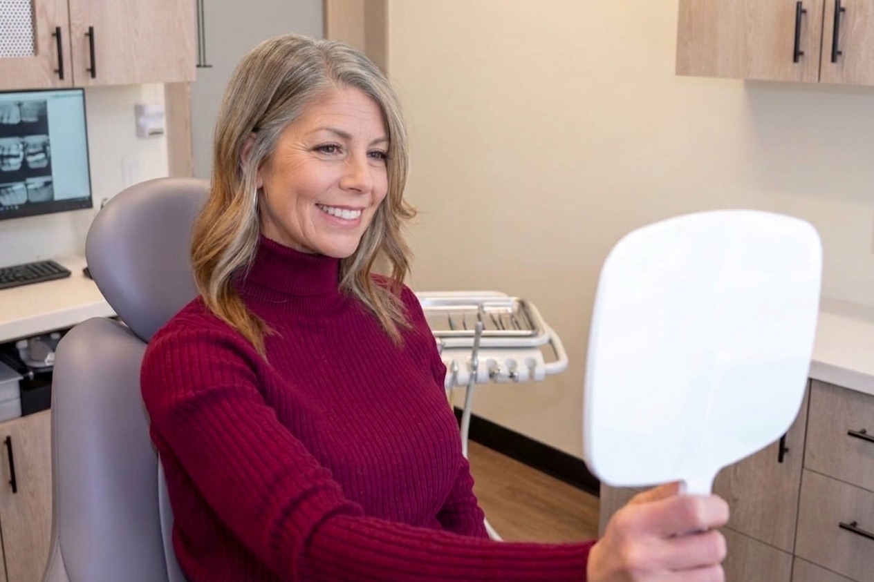 Smiling middle-aged woman with gray hair in a dental chair holding a white handheld mirror in a dental office.