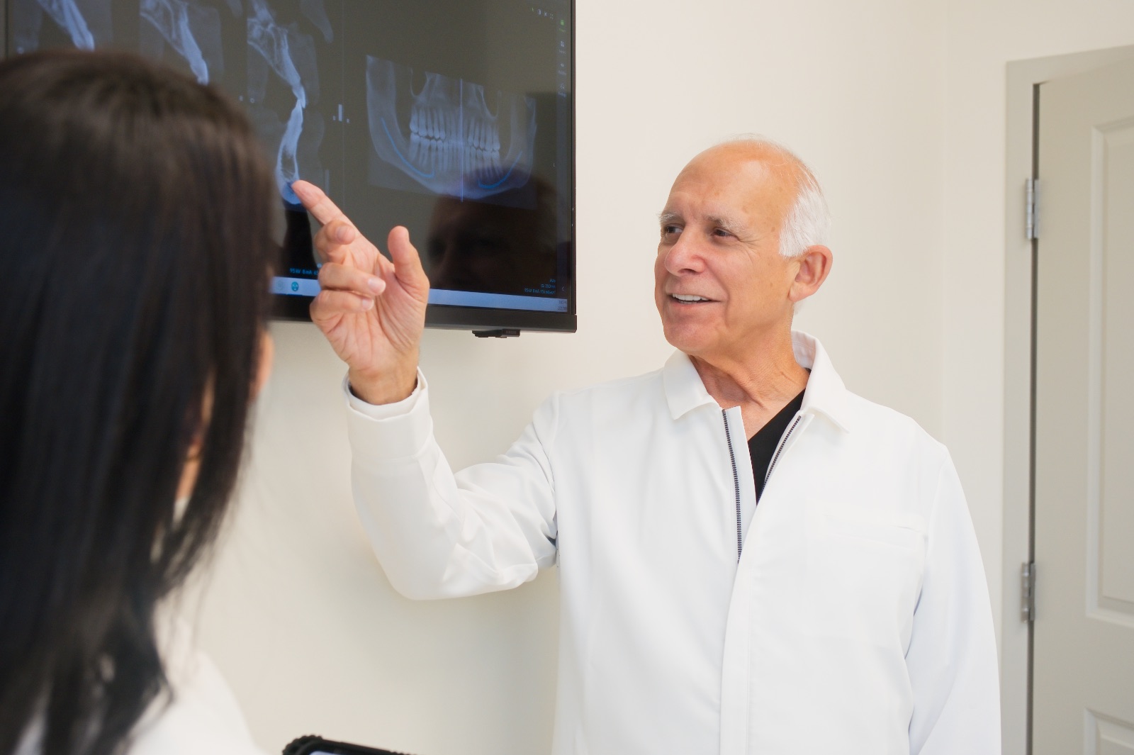 A male dentist in a white coat points to dental X-rays displayed on a screen while talking to a patient.
