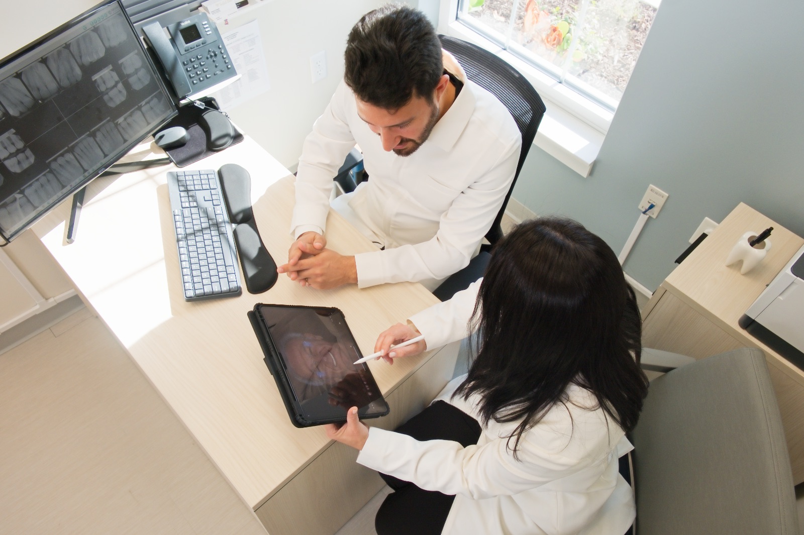 Two medical professionals in white coats sitting at a desk; one is showing the other a digital tablet with a stylus, while a computer screen displays dental X-rays.