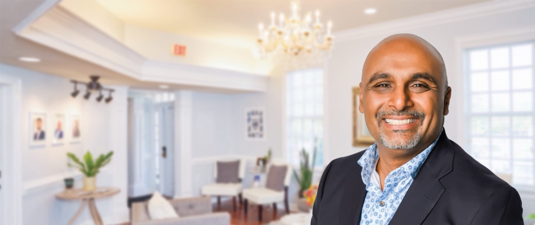 Smiling bald man with a trimmed gray beard wearing a dark blazer and blue patterned shirt in a well-lit, elegant room with white walls, large windows, and comfortable chairs.