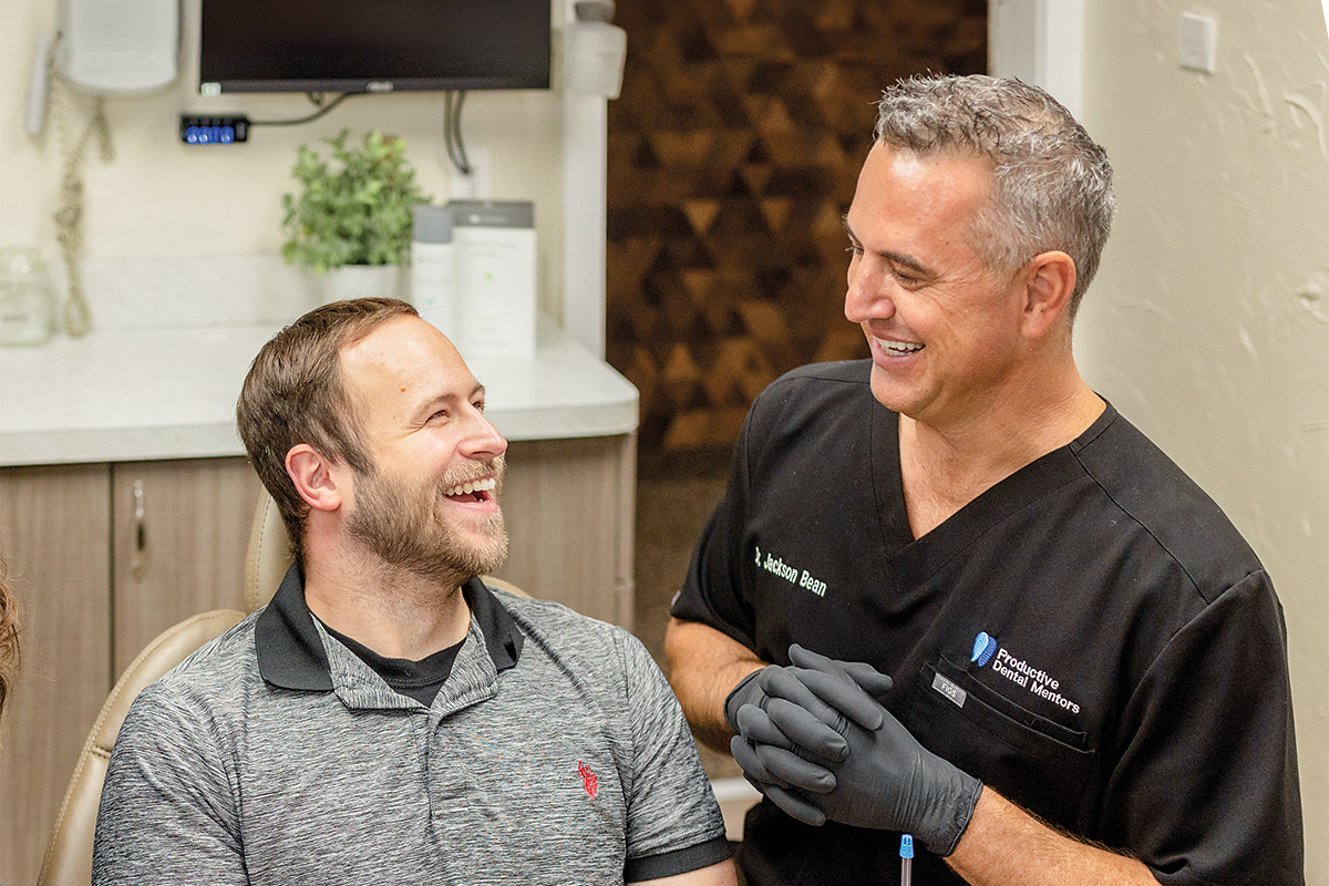 Smiling dentist in black scrubs with 'Dr. Jackson Bean' and 'Productive Dental Mentors' logos chatting with a happy male patient seated in a dental chair.