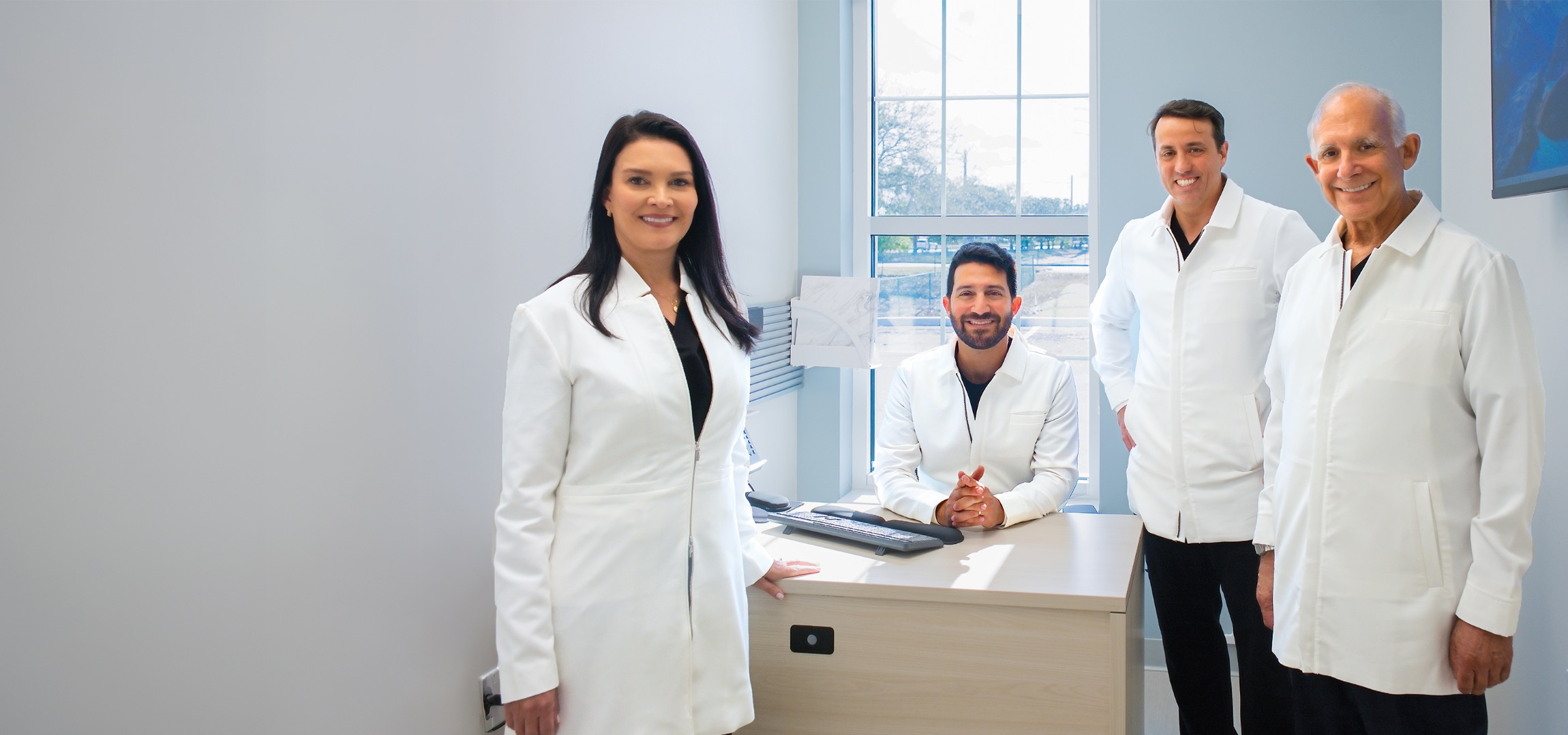 Four medical professionals wearing white lab coats in a bright office, one woman standing on the left and three men, one sitting behind a desk and two standing on the right, all smiling at the camera.