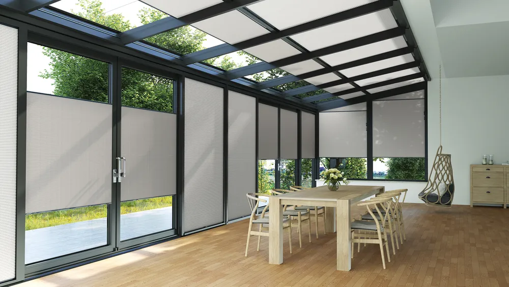 Moderner Wintergarten mit Glasdach und großen Fenstern, die mit hellen Wabenplissee-Jalousien teilweise verdunkelt sind; heller Holztisch mit sechs Stühlen und eine hängende Sitzschaukel rechts neben einer Kommode.