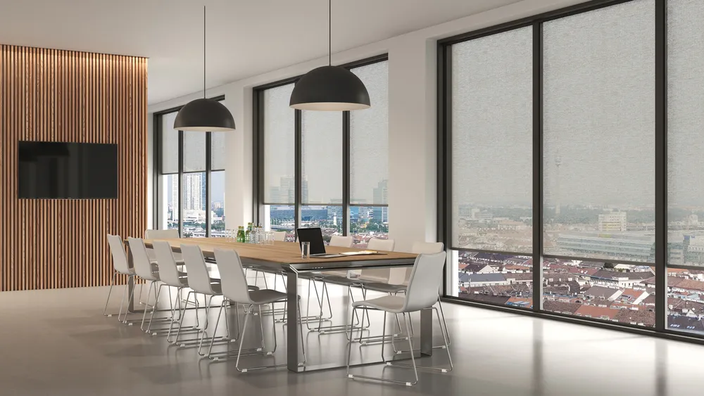 Moderner Besprechungsraum mit langem Holztisch und weißen Stühlen, großen Fenstern mit halbtransparenten Rollos und zwei schwarzen Pendelleuchten.