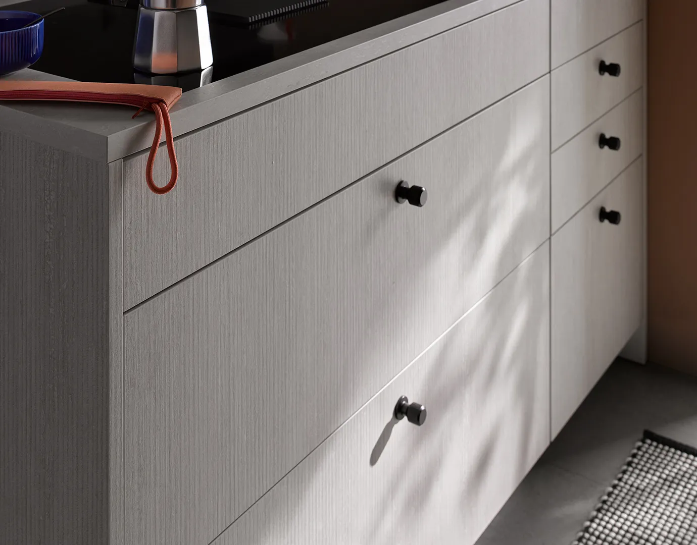 Seitenansicht einer modernen grauen Küchenkommode mit schwarzen Knöpfen, einer Edelstahl-Kaffeemaschine und einem graublauen Schüssel auf der dunklen Arbeitsfläche, mit Sonnenlicht, das Schatten auf die Kommodentür wirft.