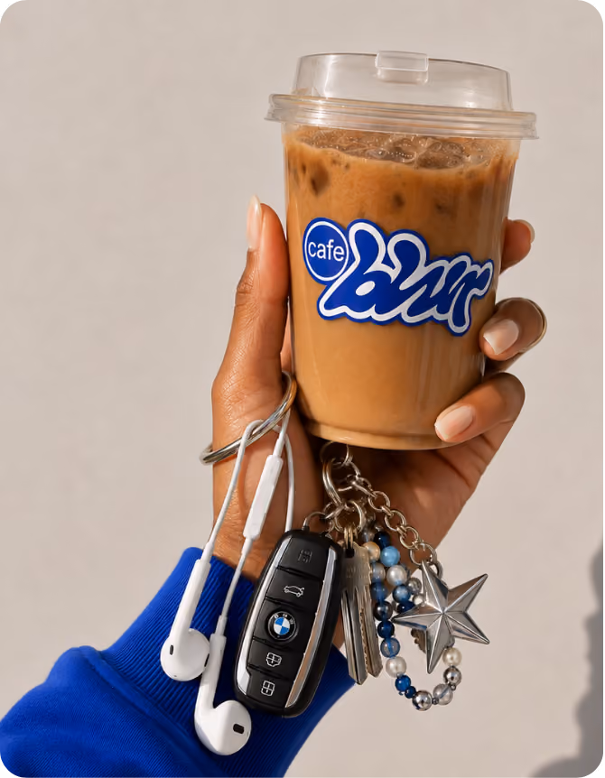 A hand wearing a blue sleeve holds a plastic cup of iced coffee with a clear lid and a blue and white 'cafe blur' logo, also holding earphones and a BMW car key attached to a keychain with blue and silver beads and a silver star charm.