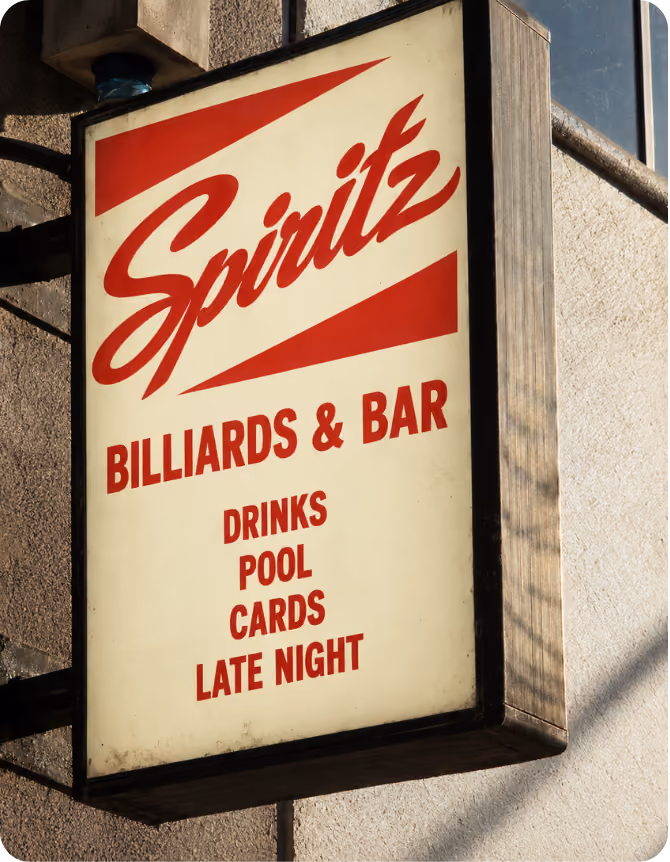 Outdoor rectangular sign on a building wall reading 'Spritz' in large red script with 'BILLIARDS & BAR' below and 'DRINKS POOL CARDS LATE NIGHT' listed underneath in red capital letters.