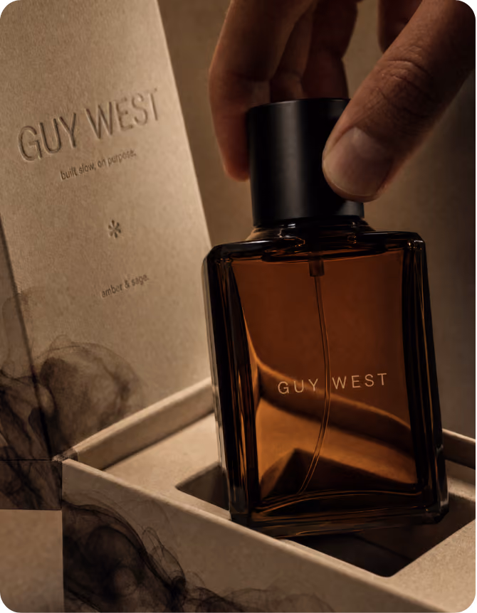 Hand holding a dark amber rectangular perfume bottle labeled 'GUY WEST' inside an open textured box that also displays 'GUY WEST built slow, on purpose amber & sage' on the inside flap.
