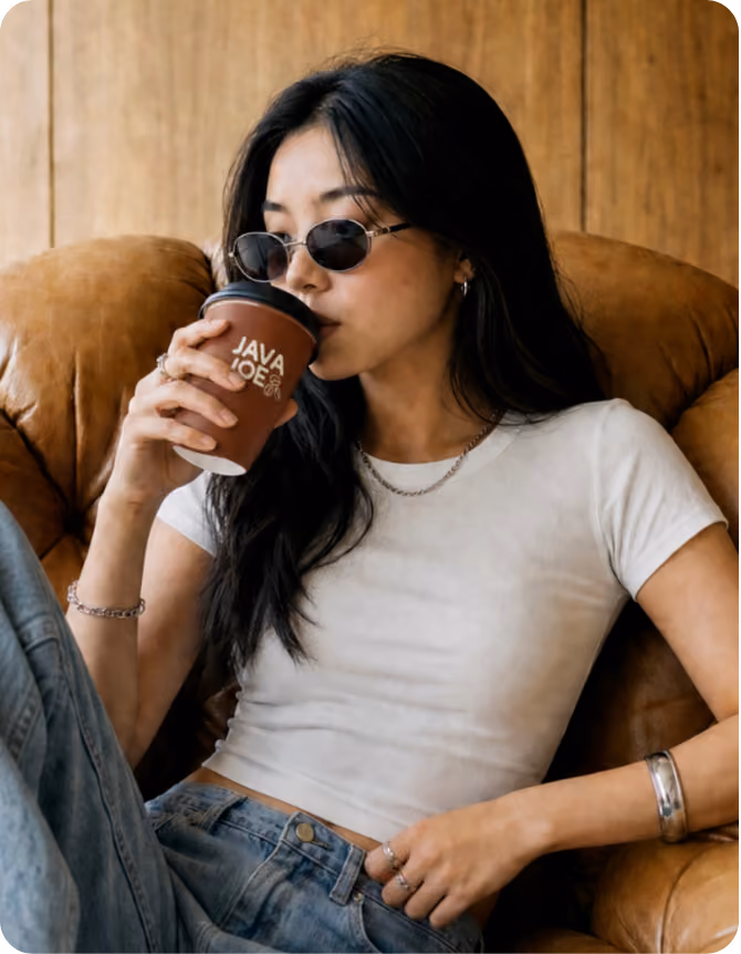 Young woman with long black hair wearing sunglasses, a white fitted t-shirt, and jeans, sitting on a brown leather chair and drinking from a brown coffee cup with the words 'JAVA JOE' on it.