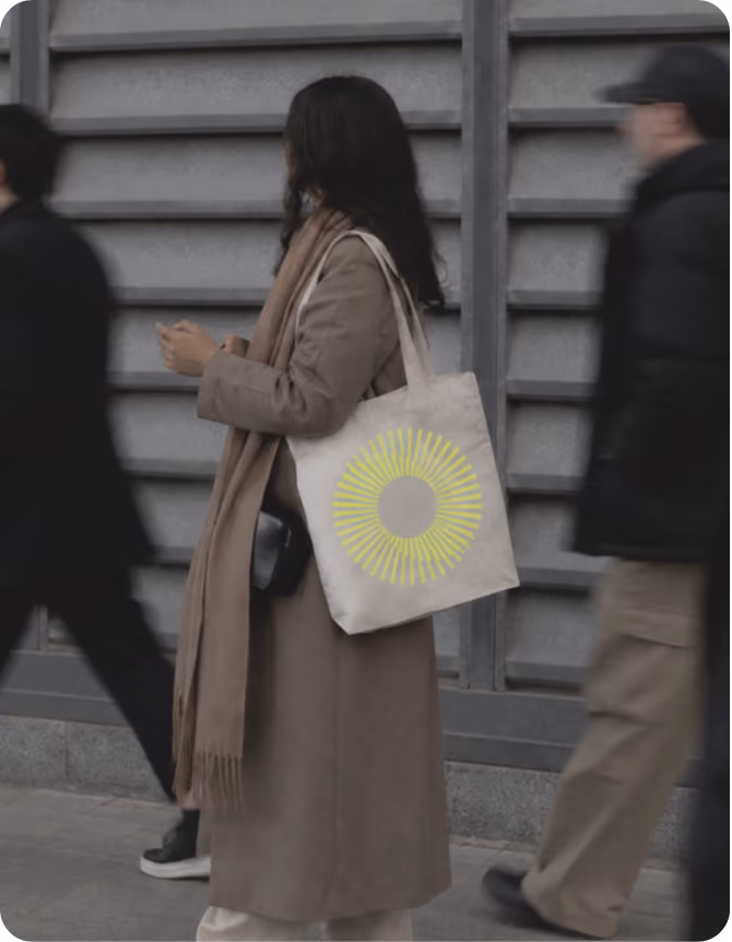 Woman in a long brown coat and scarf standing on a sidewalk holding a beige tote bag with a yellow circular sunburst design, flanked by two blurred pedestrians walking past.
