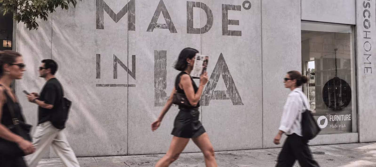 Street scene with blurred people walking past a concrete wall that has large faded text reading 'MADE IN LA'.