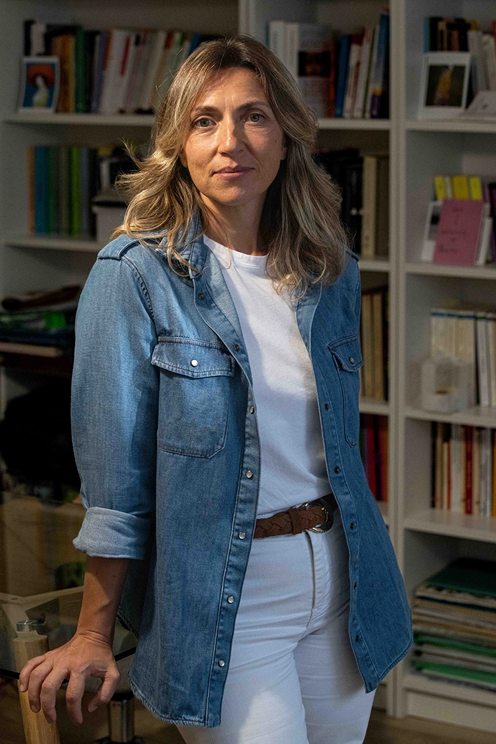 Woman with wavy blonde hair, wearing a blue denim shirt over a white t-shirt and white pants, standing in front of bookshelves filled with books and looking with empathy at the camera.