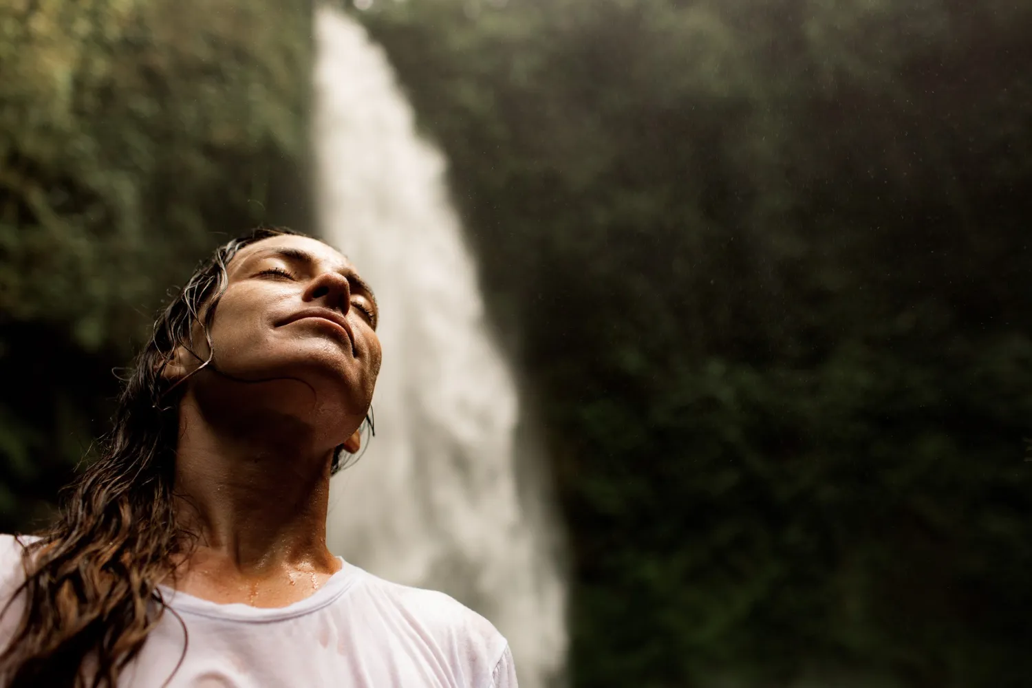 Mulher de olhos fechados junto a uma cascata, com ar sereno.