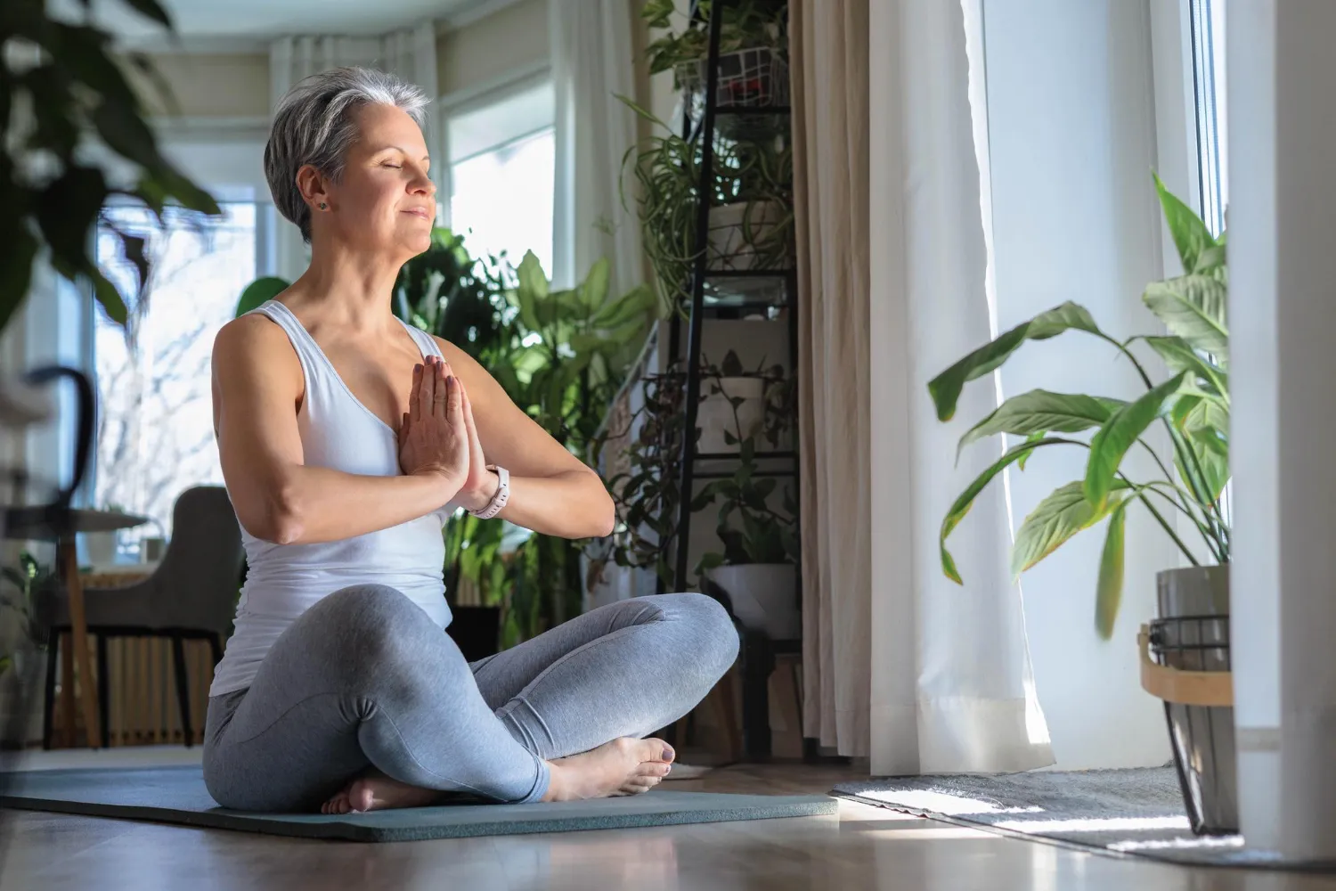Mulher de cabelo curto e grisalho a meditar sentada num tapete, de olhos fechados e mãos em prece.