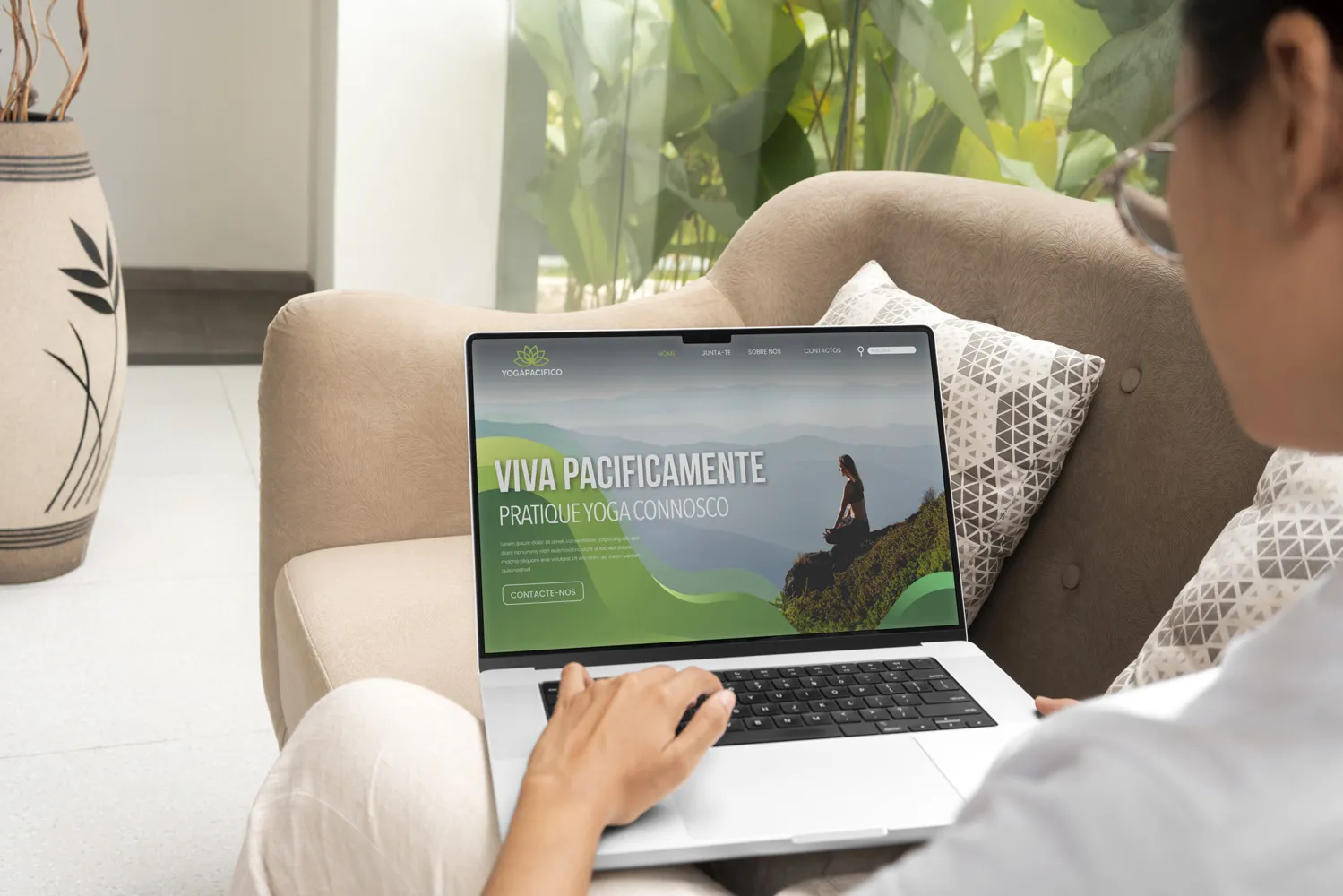 Pessoa sentada num sofá bege fazendo scroll num site de yoga no laptop com o texto 'Viva Pacificamente, pratique yoga connosco' com imagem de uma pessoa a meditar num monte com fundo de montanhas e céu nublado.