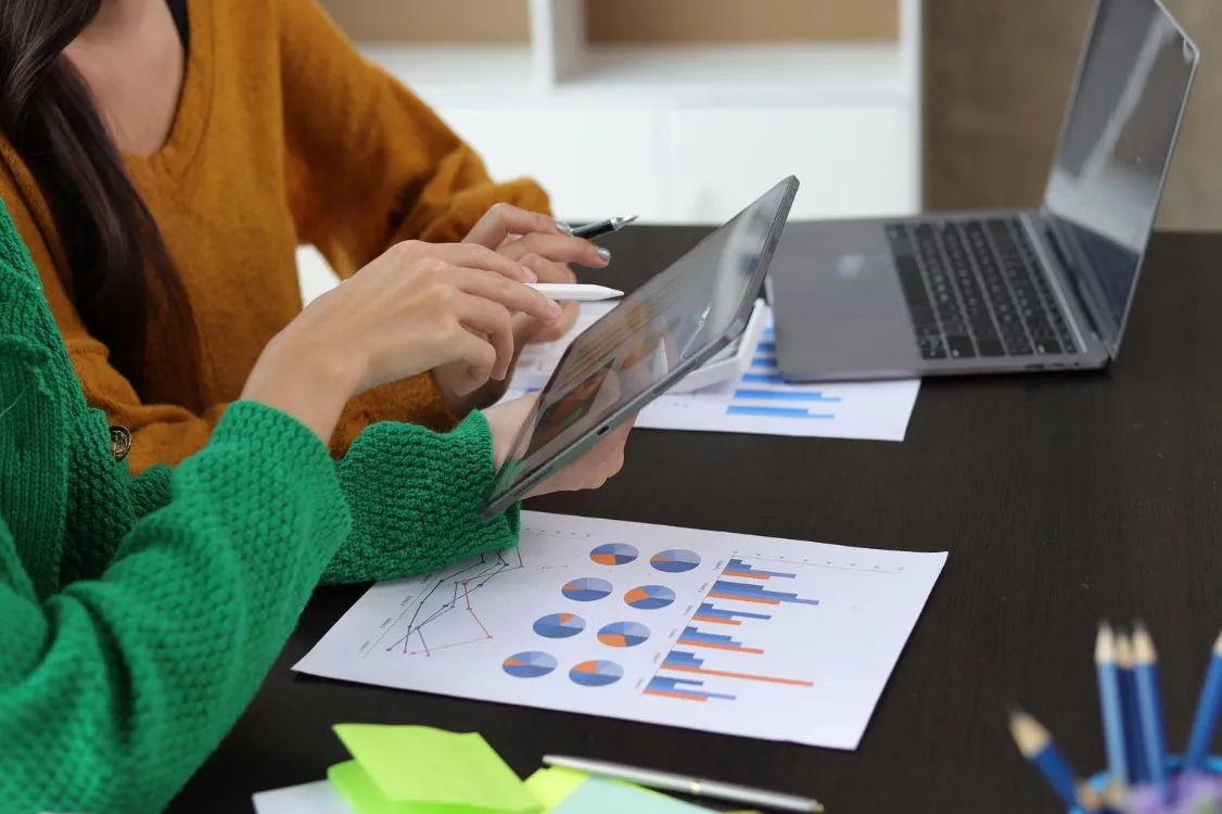 Duas pessoas colaboram usando um tablet e analisando gráficos e dados impressos em folhas na mesa, com um computador portátil aberto próximo.
