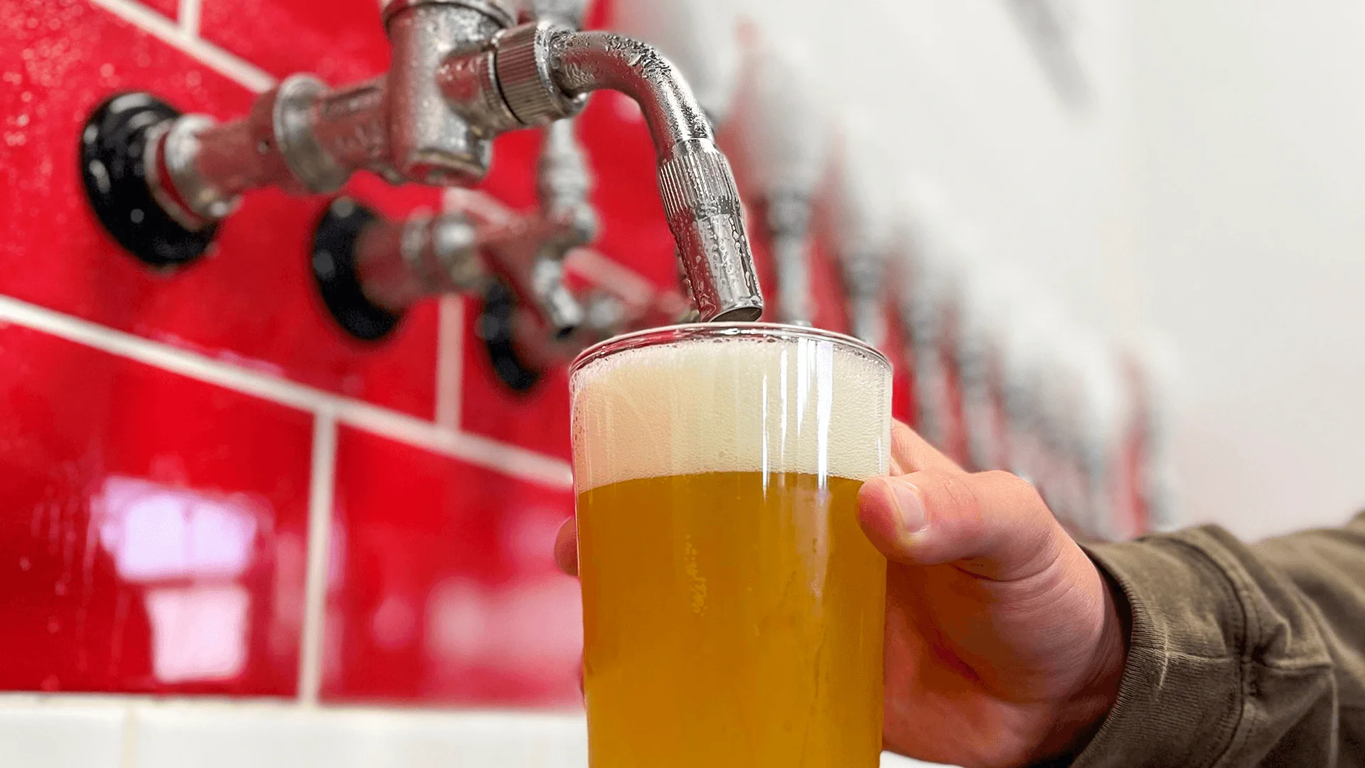 A hand holds a pint glass beneath a metal tap as golden craft beer pours in with thick foam.
