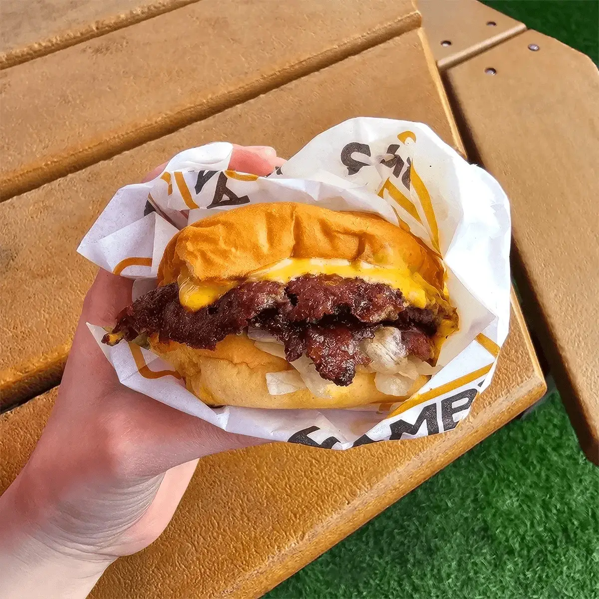 A hand holds a hot Hamby's smash burger with melted cheese and lettuce in branded wrap on an outdoor picnic table.