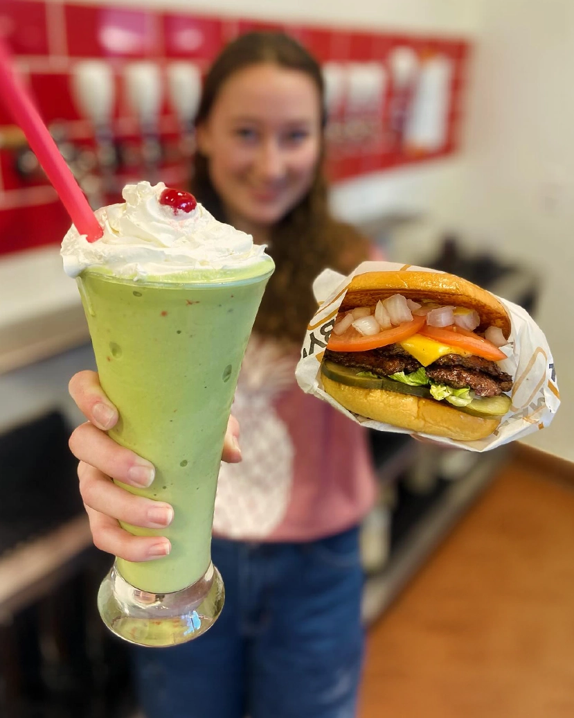 A teenage girl holds a freshly-made milkshake and smashburger, smiling at the camera.