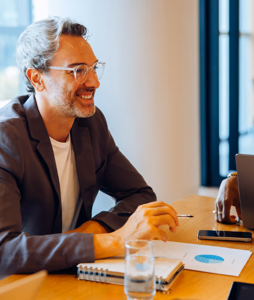 Smiling man with glasses seated at table with notebook, pen, and smartphone during meeting.