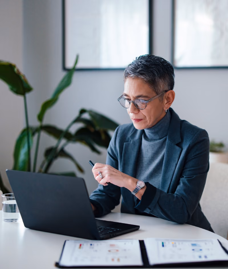 Woman in glasses and blazer working on a laptop at a desk with documents and a glass of water.
