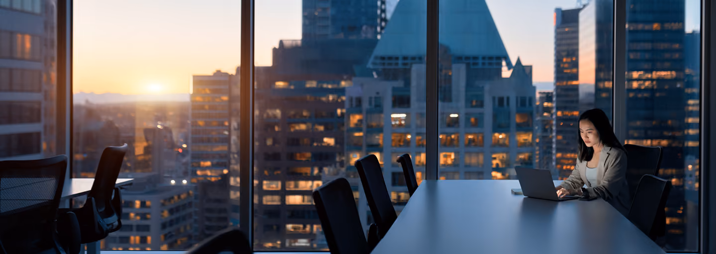 Woman working on laptop alone in a conference room with city skyline at sunset.